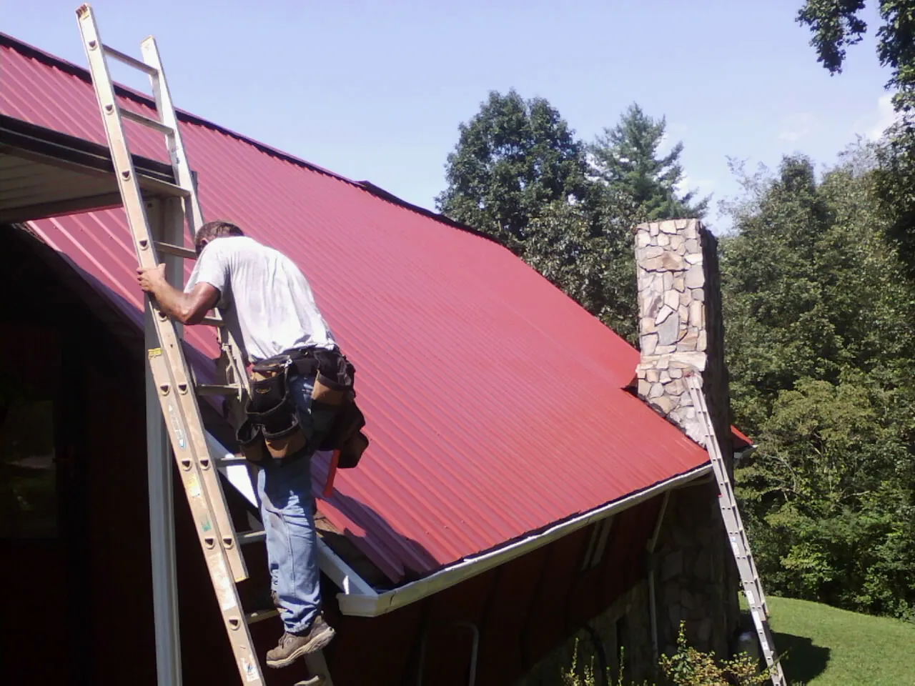 Roofer on a ladder beside a red metal roof and stone chimney — Soffit & Fascia Repair by Roofing Brothers in South Grafton